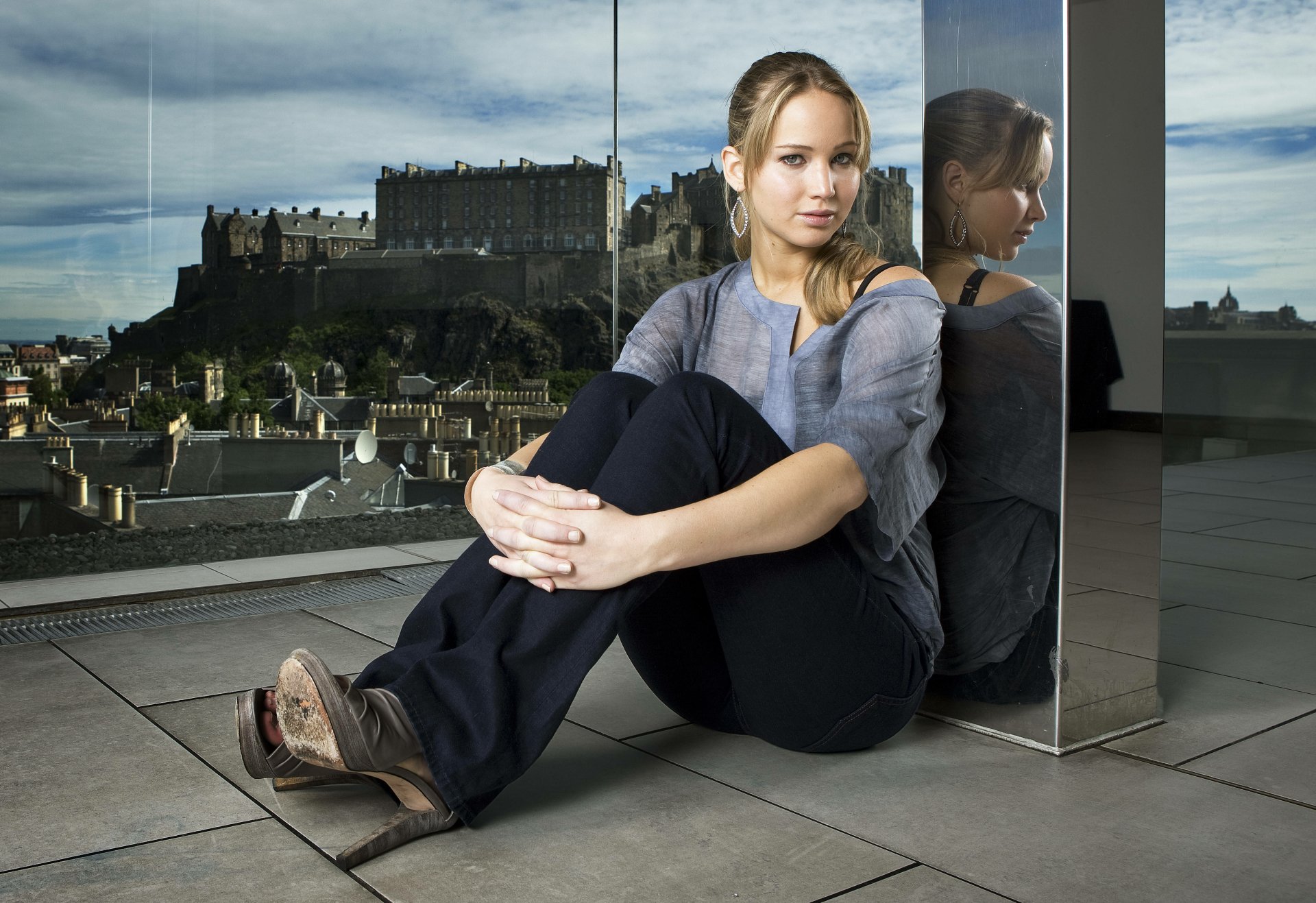 A 4K Ultra HD wallpaper featuring Jennifer Lawrence seated against a glass panel, with a view of a castle in the background and a reflective surface beside her.