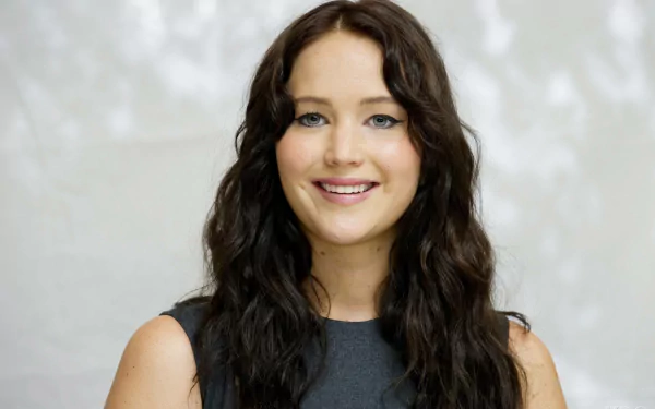 A close-up of Jennifer Lawrence smiling, featuring her long, wavy hair and wearing a simple black outfit, set against a soft, blurred background. This image serves as an HD desktop wallpaper.