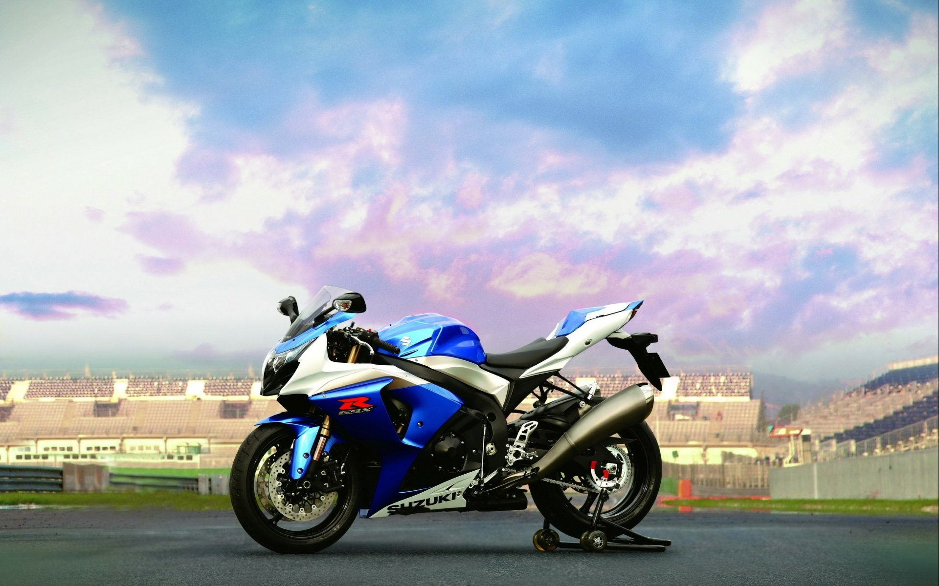 Blue Suzuki GSX-R1000 sportbike on a racetrack under a dramatic sky, 2K Quad HD PC desktop wallpaper background featuring a Suzuki vehicle.