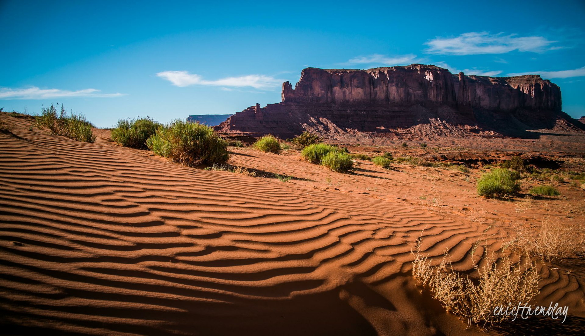 HD PC desktop wallpaper and background: Monument Valley, Arizona desert nature scene — rippled sand, sparse shrubs and a distant butte beneath a bright blue sky.