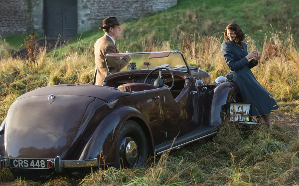 A scene from the TV show Outlander, featuring two characters by a vintage convertible car in a grassy field, capturing a moment of connection amidst scenic surroundings.