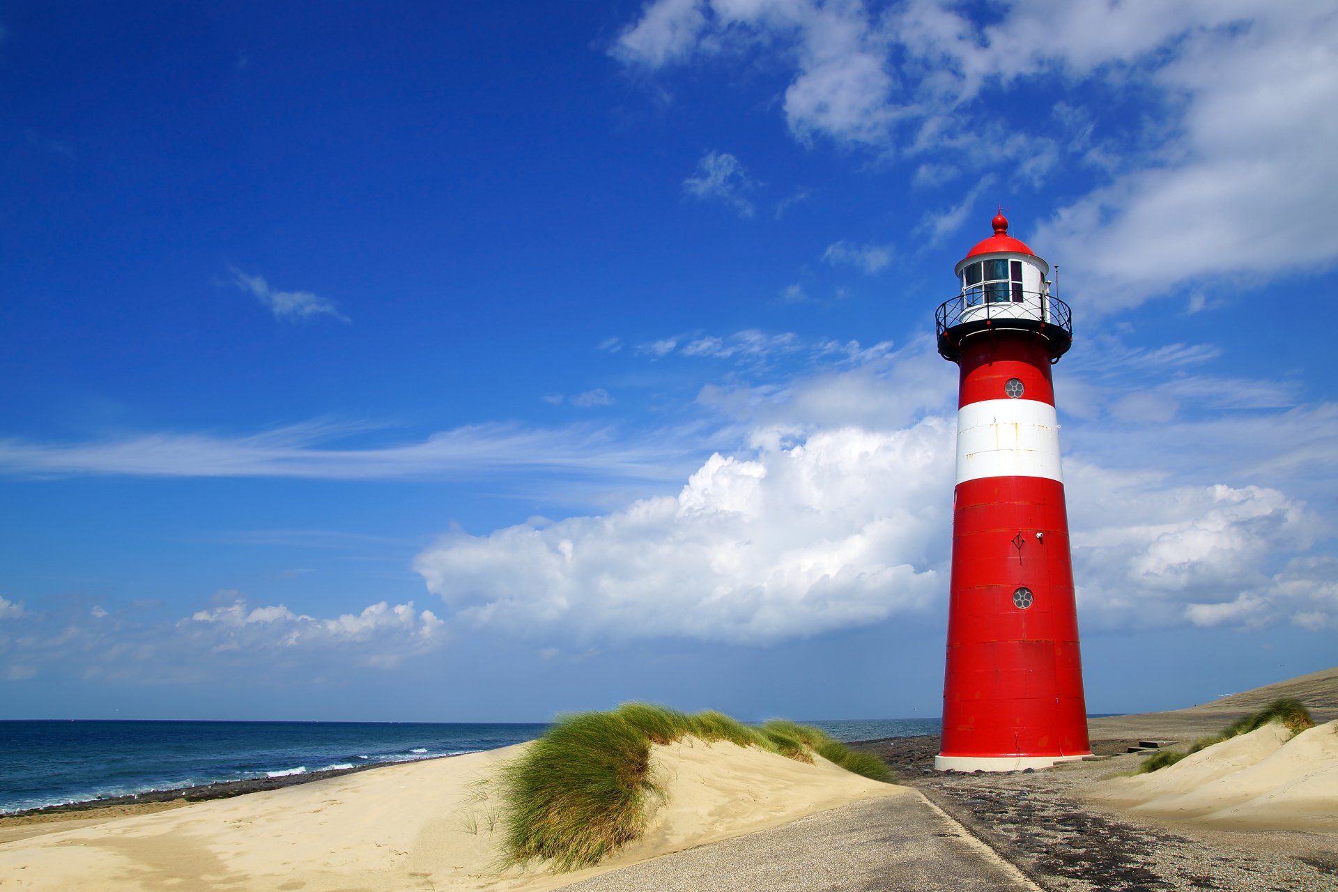 4K Ultra HD wallpaper of a red and white man-made lighthouse standing on sandy dunes under a vibrant blue sky with scattered clouds by the ocean.