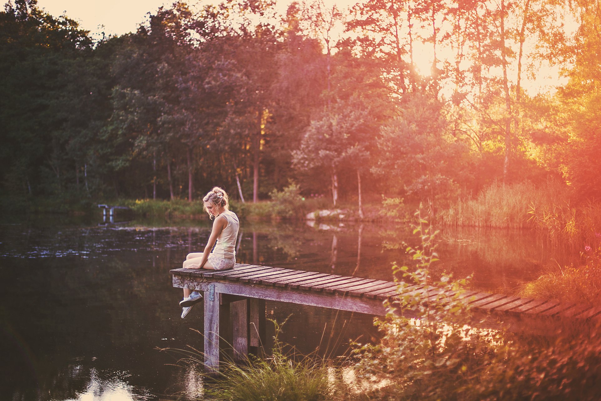 A serene HD desktop wallpaper featuring a woman sitting at the edge of a wooden dock by a tranquil lake, surrounded by lush trees and bathed in warm, golden sunlight.