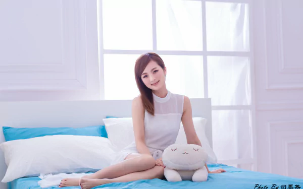 Taiwanese woman in a white dress smiling while sitting on a bed with white pillows and a plush toy, captured in a bright, airy room.