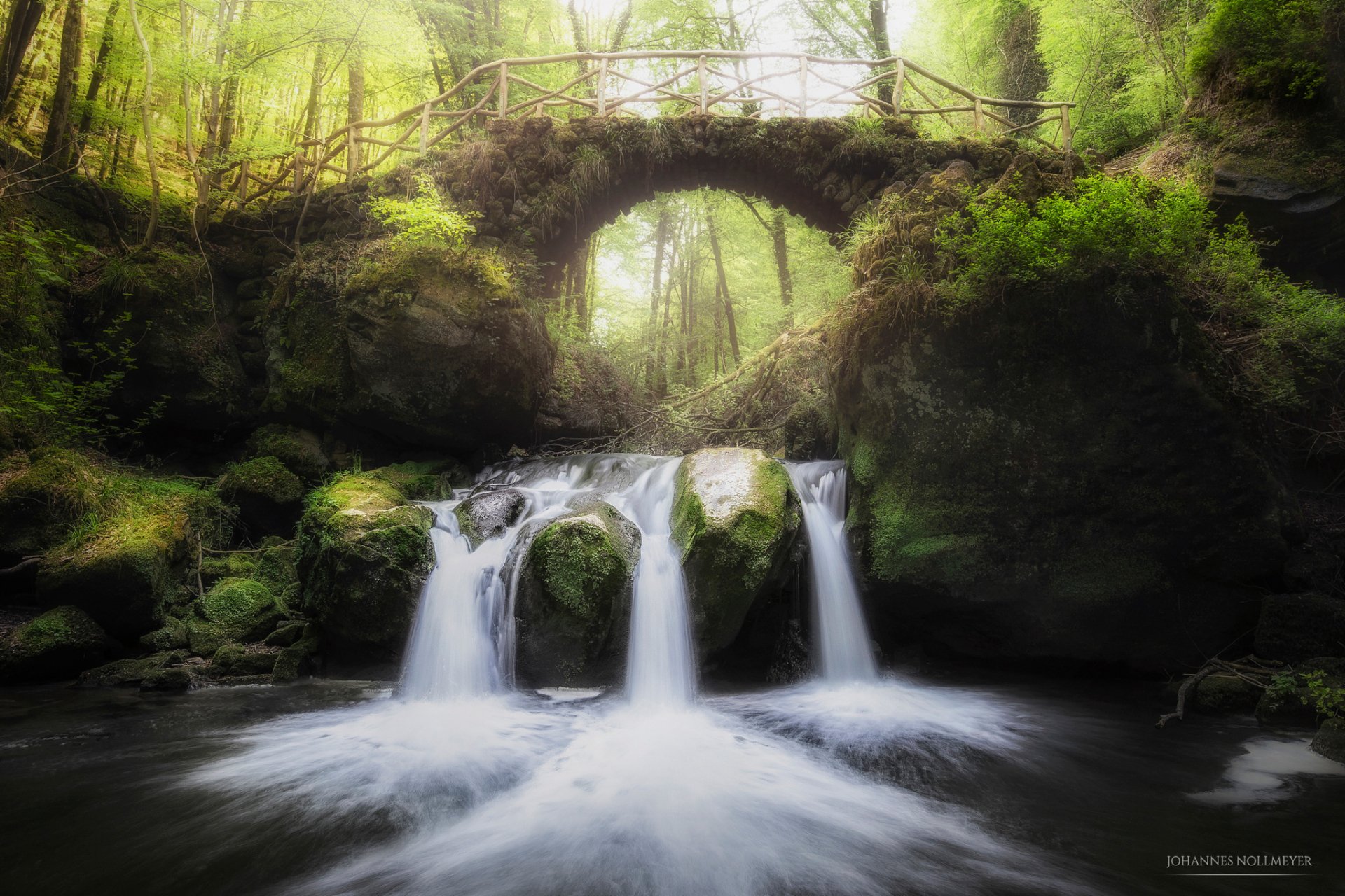 A serene HD desktop wallpaper of Muellerthal, Luxembourg, featuring a lush forest landscape with a cascading waterfall beneath an old stone bridge.