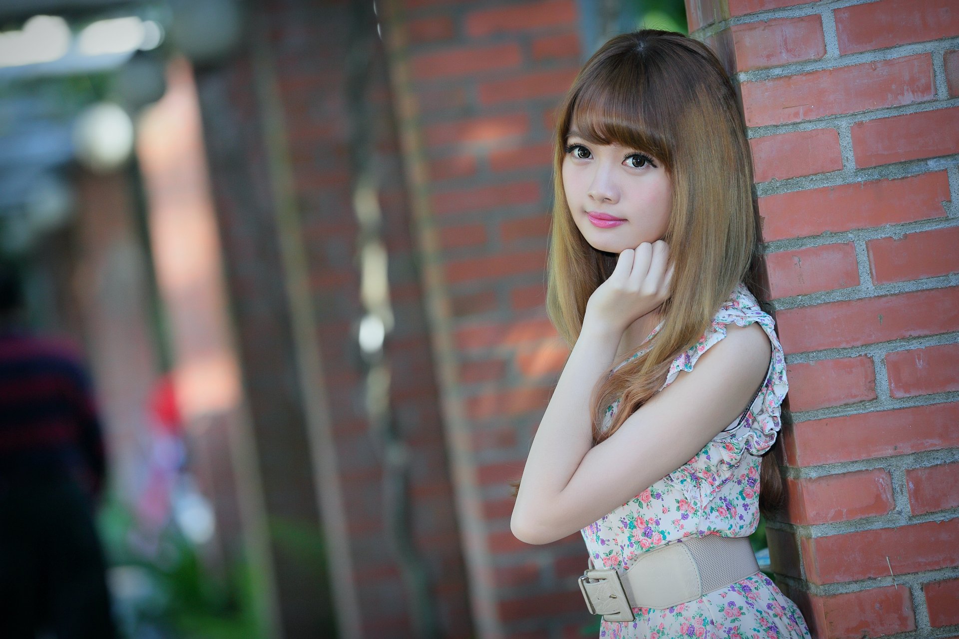A Taiwanese model poses elegantly by a brick wall, wearing a floral dress and a belt, with soft bokeh in the background. The image captures her beauty in vibrant 4K Ultra HD.