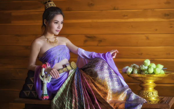 A woman in a vibrant traditional costume adorned with a belt and necklace sits gracefully, showcasing her hairstyle. The background features natural elements, enhancing the cultural elegance.
