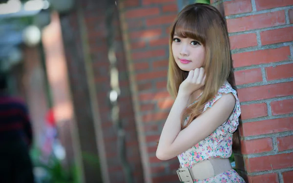 A Taiwanese model poses elegantly by a brick wall, wearing a floral dress and a belt, with soft bokeh in the background. The image captures her beauty in vibrant 4K Ultra HD.