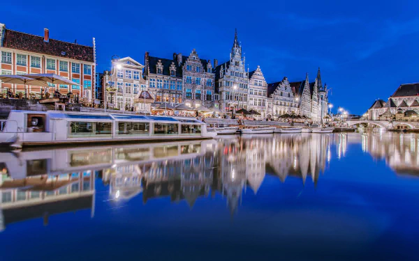 A stunning 4K Ultra HD evening view of the Bruges harbor in Belgium, showcasing boats on the river with historic city buildings reflected in the calm sea.