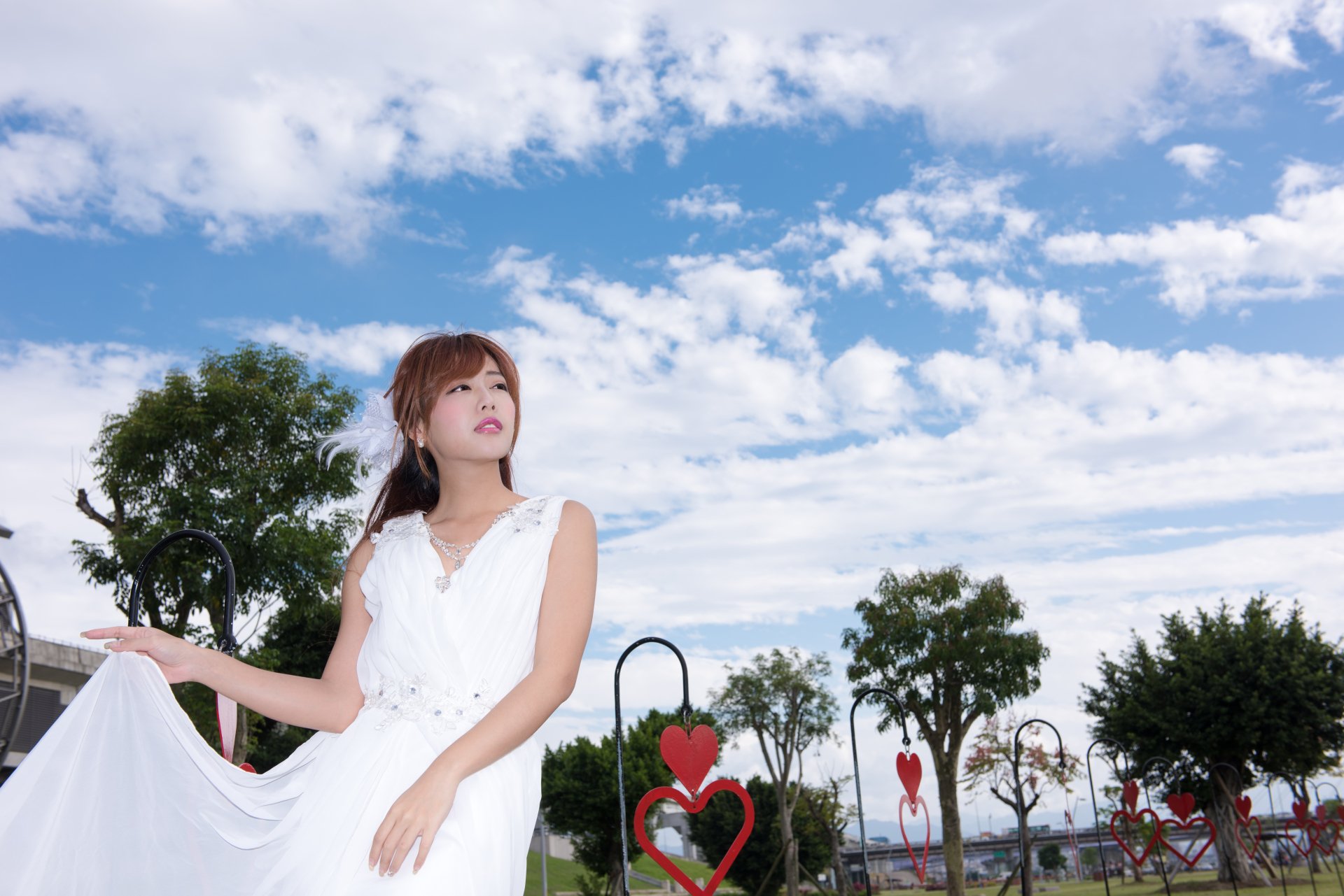 Model in a flowing white dress beneath a bright blue sky with puffy clouds, posing near heart-shaped park sculptures — 5K Ultra HD desktop wallpaper.