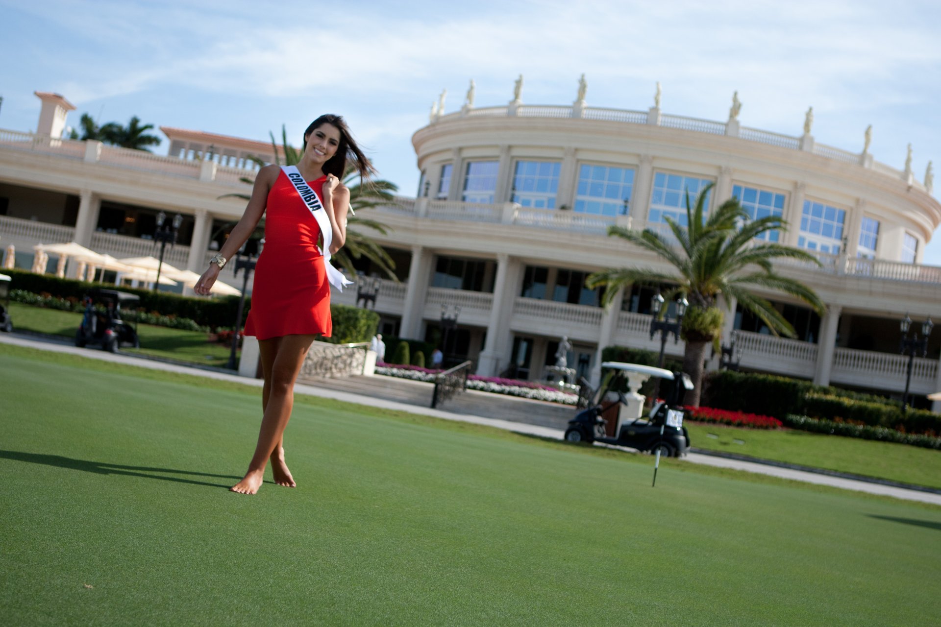 A smiling Paulina Vega, Miss Universe and Miss Colombia, poses in a vibrant red dress on a lush green lawn, with an elegant building and palm trees in the background.