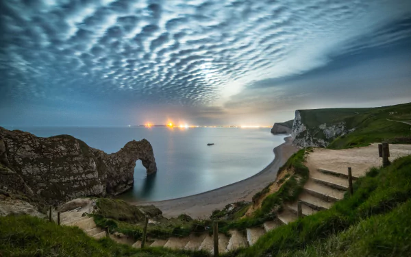 Limestone arch of Durdle Door on Dorset's coastline, England, under a textured cloud sky with calm sea and green cliffs along the shore at dusk.