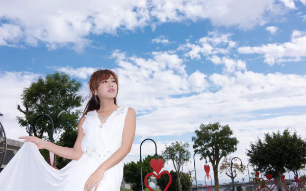 Model in a flowing white dress beneath a bright blue sky with puffy clouds, posing near heart-shaped park sculptures — 5K Ultra HD desktop wallpaper.