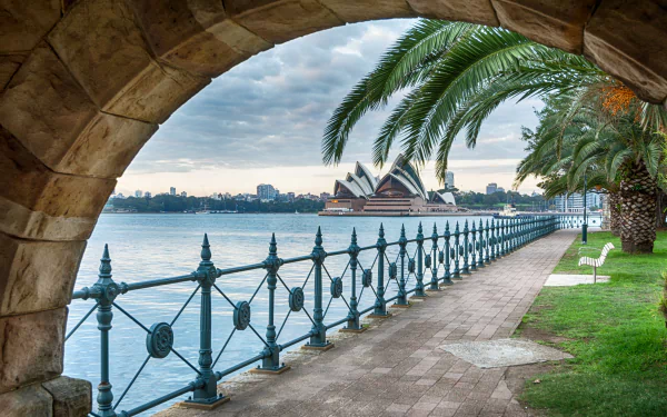 Stone arch framing a palm-lined Sydney Harbour promenade, with the man-made sails of the Sydney Opera House across the water — 5K Ultra HD desktop wallpaper, Australia.