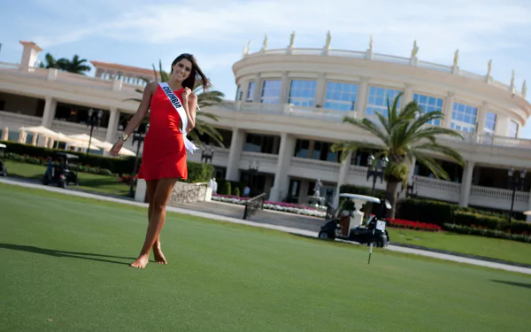 A smiling Paulina Vega, Miss Universe and Miss Colombia, poses in a vibrant red dress on a lush green lawn, with an elegant building and palm trees in the background.