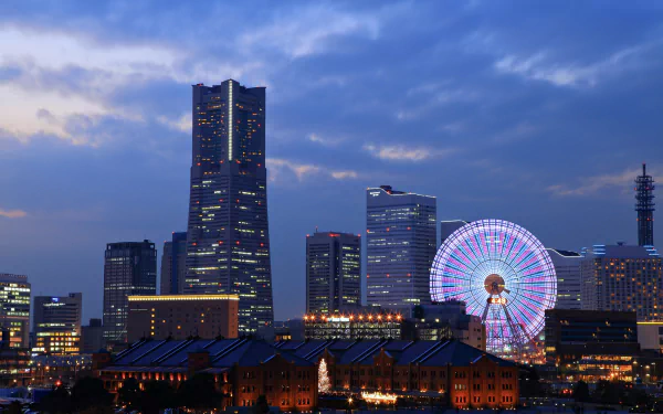 Night view of Yokohama city skyline in Japan featuring illuminated skyscrapers and a brightly lit ferris wheel against a deep blue evening sky.
