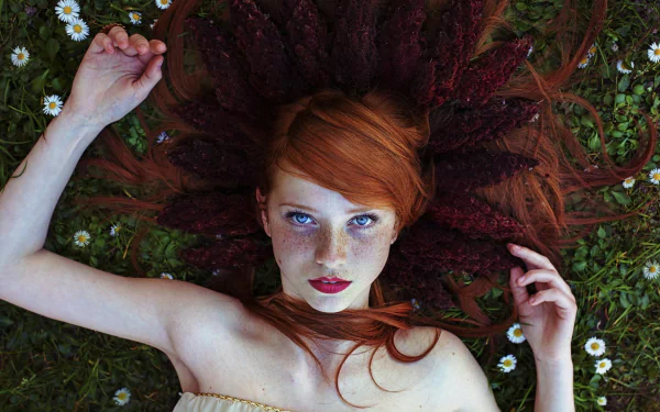 A redhead woman with freckles lies in grass, surrounded by daisies, her long hair arranged like a crown. She gazes at the viewer with striking blue eyes, evoking a serene and natural beauty.