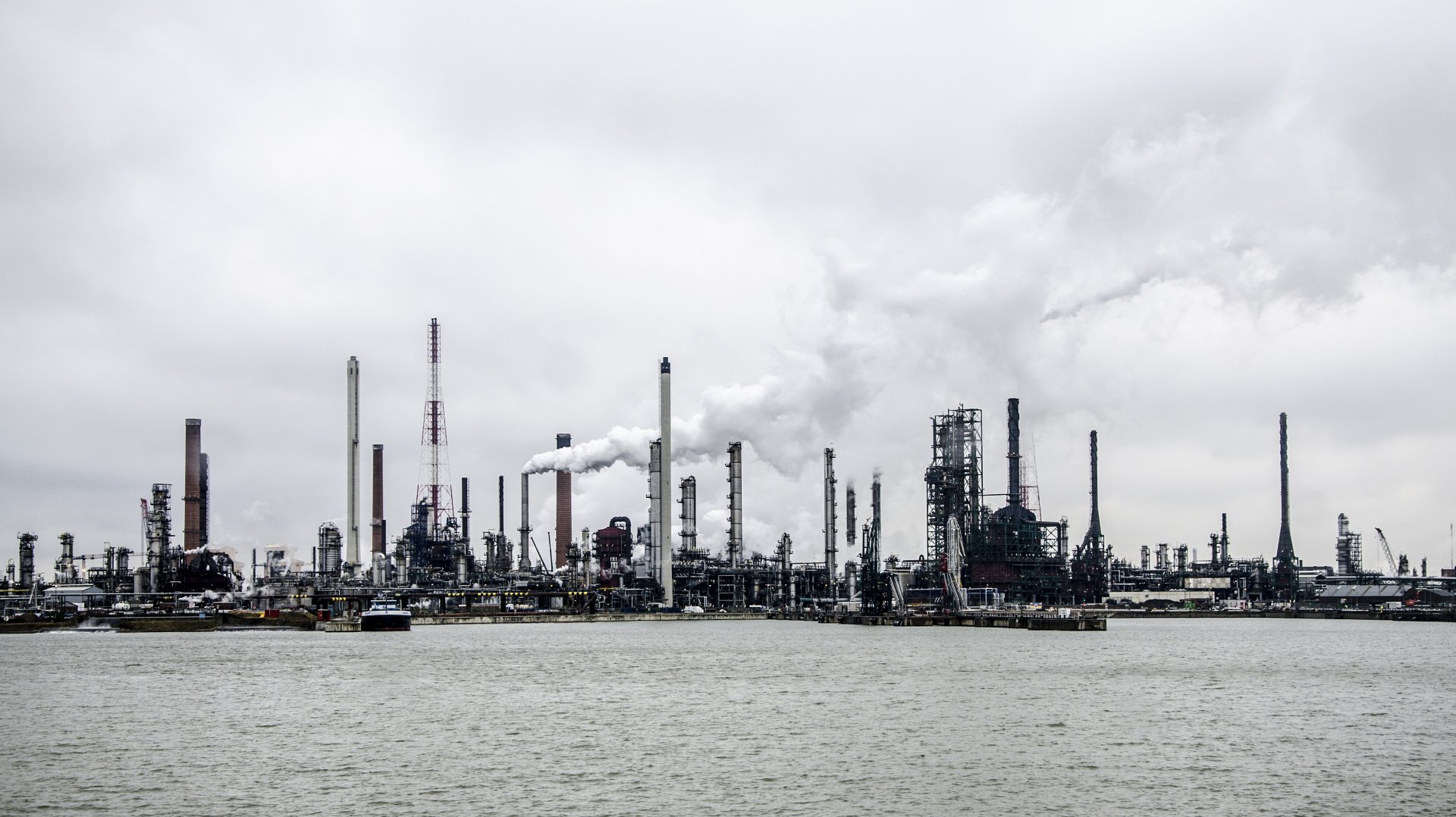 4K Ultra HD desktop wallpaper of an expansive industrial refinery complex with smokestacks and pipes under a cloudy sky by the water.