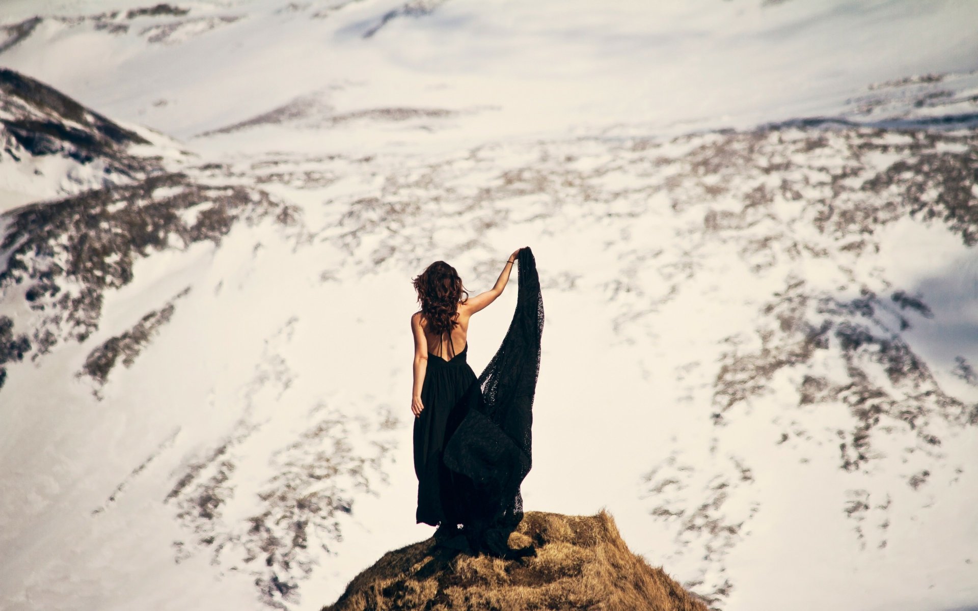 A brunette woman in a flowing black dress stands on a rocky peak overlooking a vast snowy mountain landscape, captured in 4K Ultra HD for a desktop wallpaper.