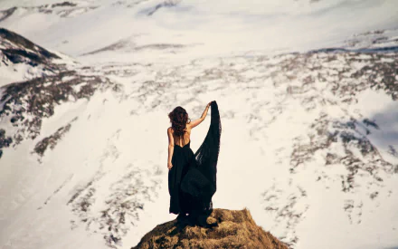 A brunette woman in a flowing black dress stands on a rocky peak overlooking a vast snowy mountain landscape, captured in 4K Ultra HD for a desktop wallpaper.