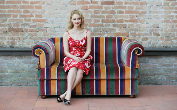 Canadian actress Sarah Gadon with blonde hair and blue eyes sits elegantly on a colorful striped sofa against a brick wall background.
