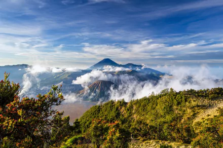 A breathtaking view of Mount Bromo, a stratovolcano in Java, Indonesia, surrounded by lush greenery and clouds under a vibrant sky, making an impressive nature landscape.