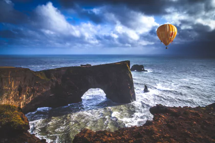 5K Ultra HD landscape photograph of Dyrhólaey sea arch in Iceland, turbulent sea below and a yellow hot air balloon drifting above — seascape desktop wallpaper.