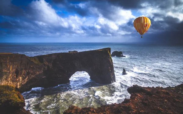 5K Ultra HD landscape photograph of Dyrhólaey sea arch in Iceland, turbulent sea below and a yellow hot air balloon drifting above — seascape desktop wallpaper.