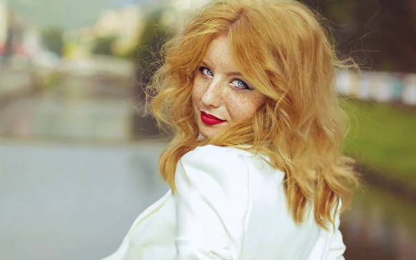 A redhead woman with blue eyes and freckles smiles at the camera, showcasing vibrant red lipstick. The image serves as a stunning HD wallpaper, blending beauty and elegance.