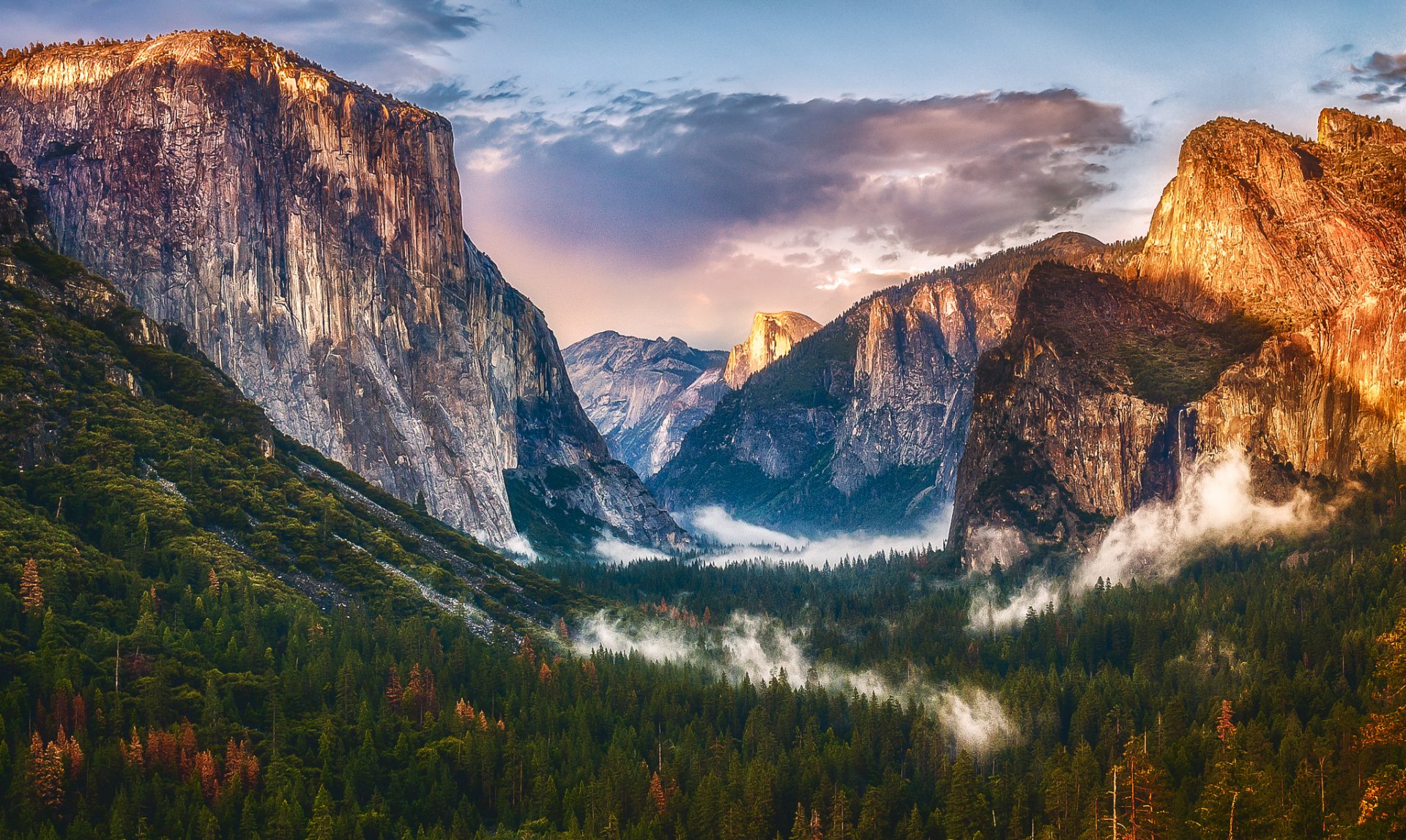HD desktop wallpaper of a dramatic mountain landscape with towering cliffs, a lush forest valley, and mist weaving through the natural scene at sunrise.