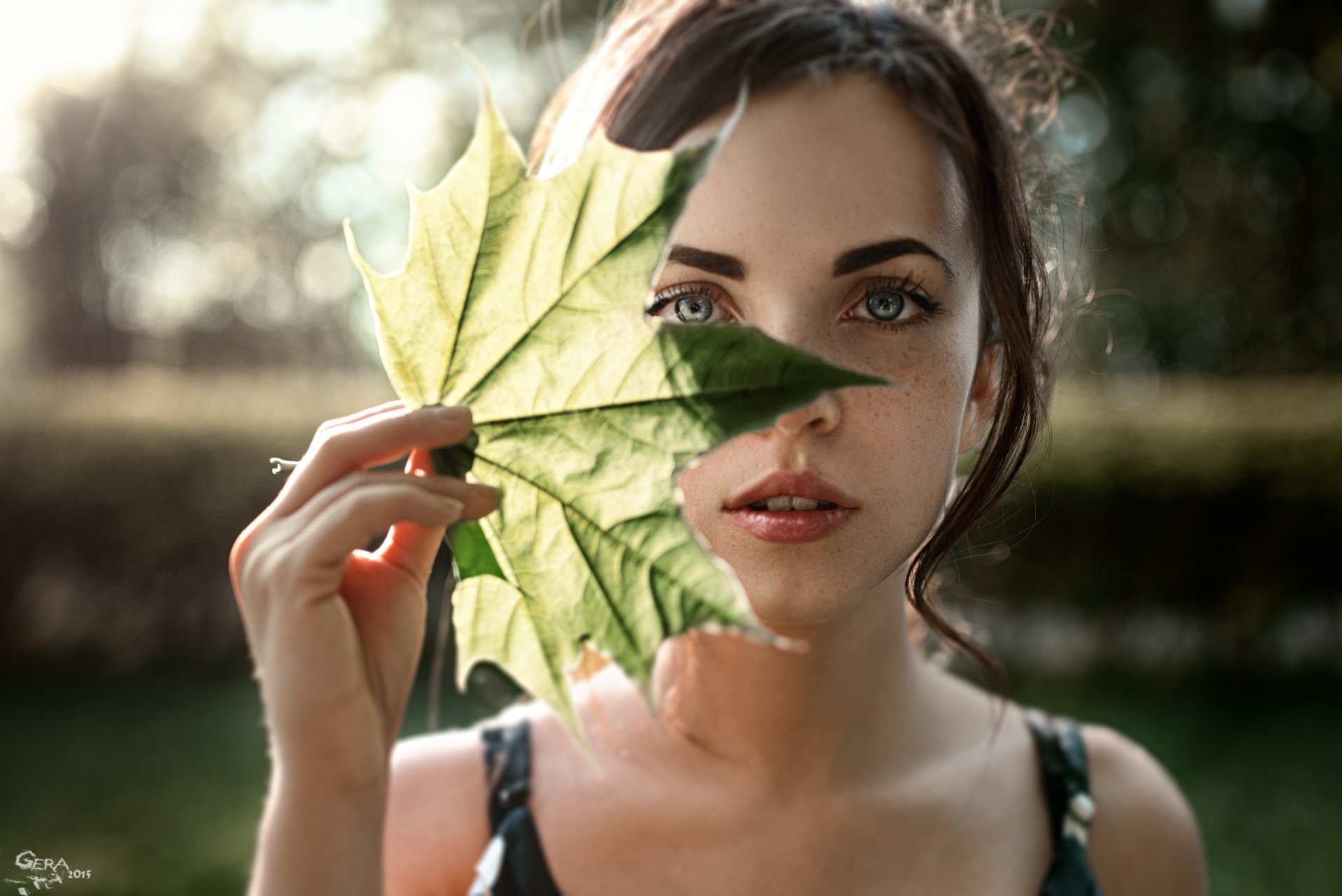 A brunette woman with blue eyes and freckles holds a green leaf to her face, surrounded by a sunny backdrop of nature, showcasing a serene and vibrant portrait.