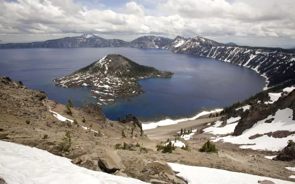 A scenic view of Crater Lake in Crater Lake National Park, Utah, surrounded by rugged snow-dusted mountains under a cloudy sky.