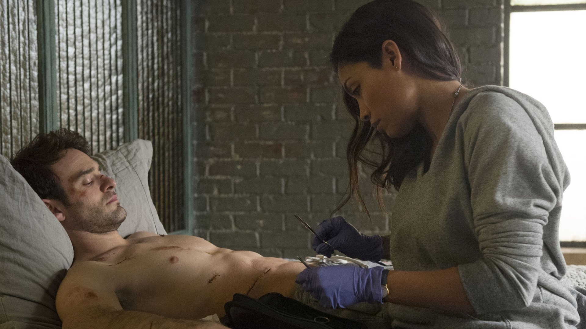 Rosario Dawson as Claire Temple from the TV show Daredevil tending to a shirtless man in a dimly lit room, captured in a high-definition desktop wallpaper.