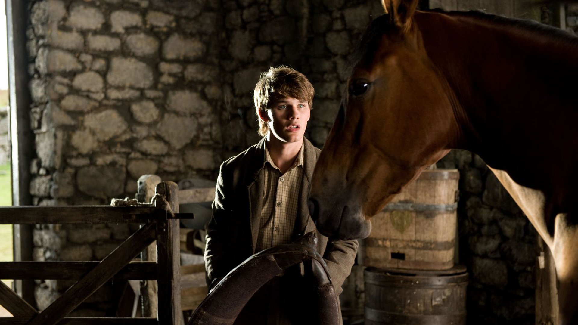 HD desktop wallpaper featuring a scene from the movie War Horse, showing a young man and a horse inside a rustic stone stable.