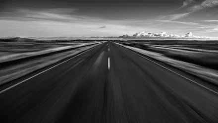 A black and white landscape image featuring a wide, straight road stretching towards the horizon, with motion blur effects and dramatic clouds in the background.