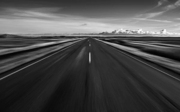 A black and white landscape image featuring a wide, straight road stretching towards the horizon, with motion blur effects and dramatic clouds in the background.