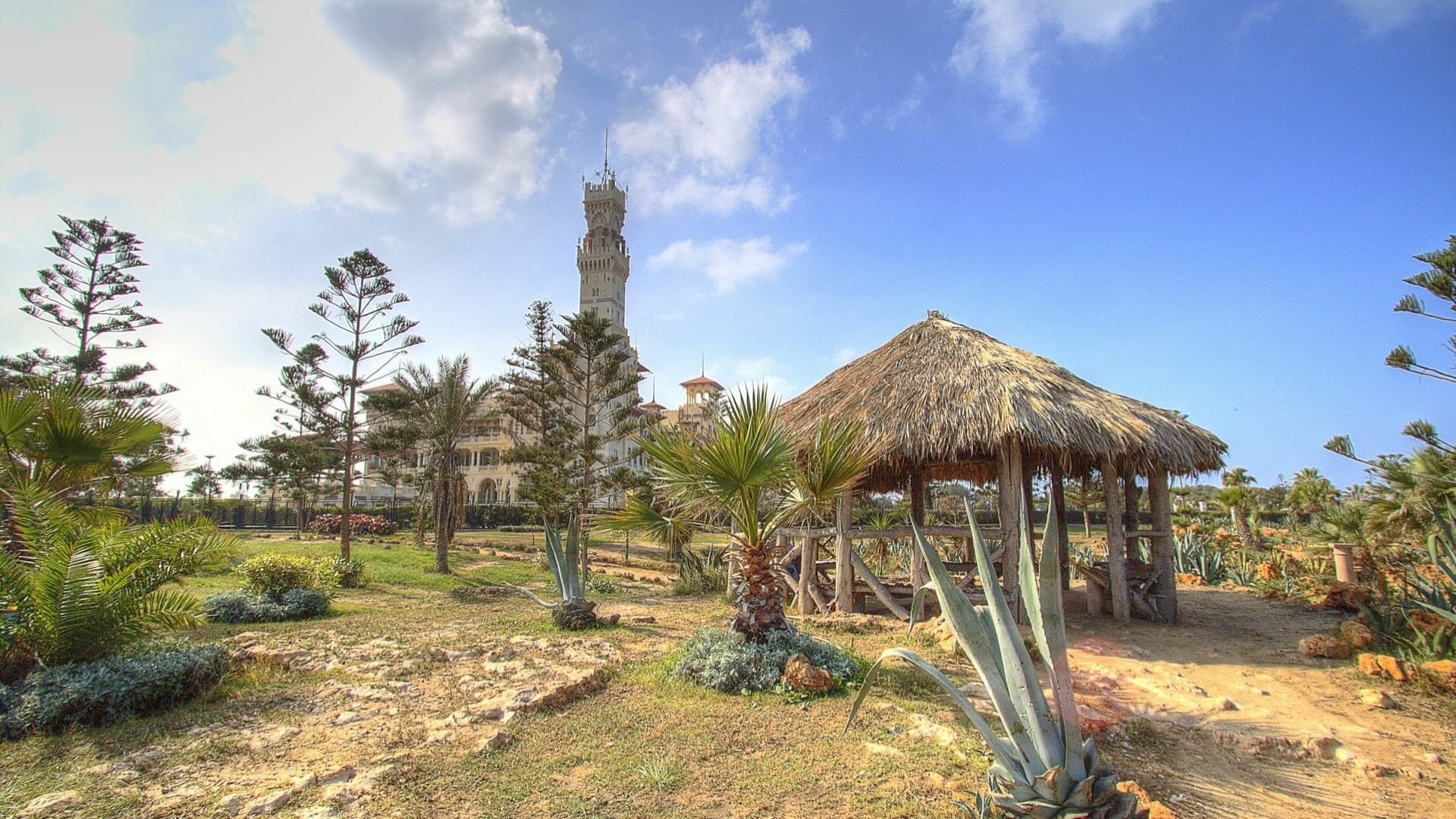Man-made Montaza Palace framed by gardens, palms and a thatched pavilion under a bright blue sky — HD PC desktop wallpaper/background.