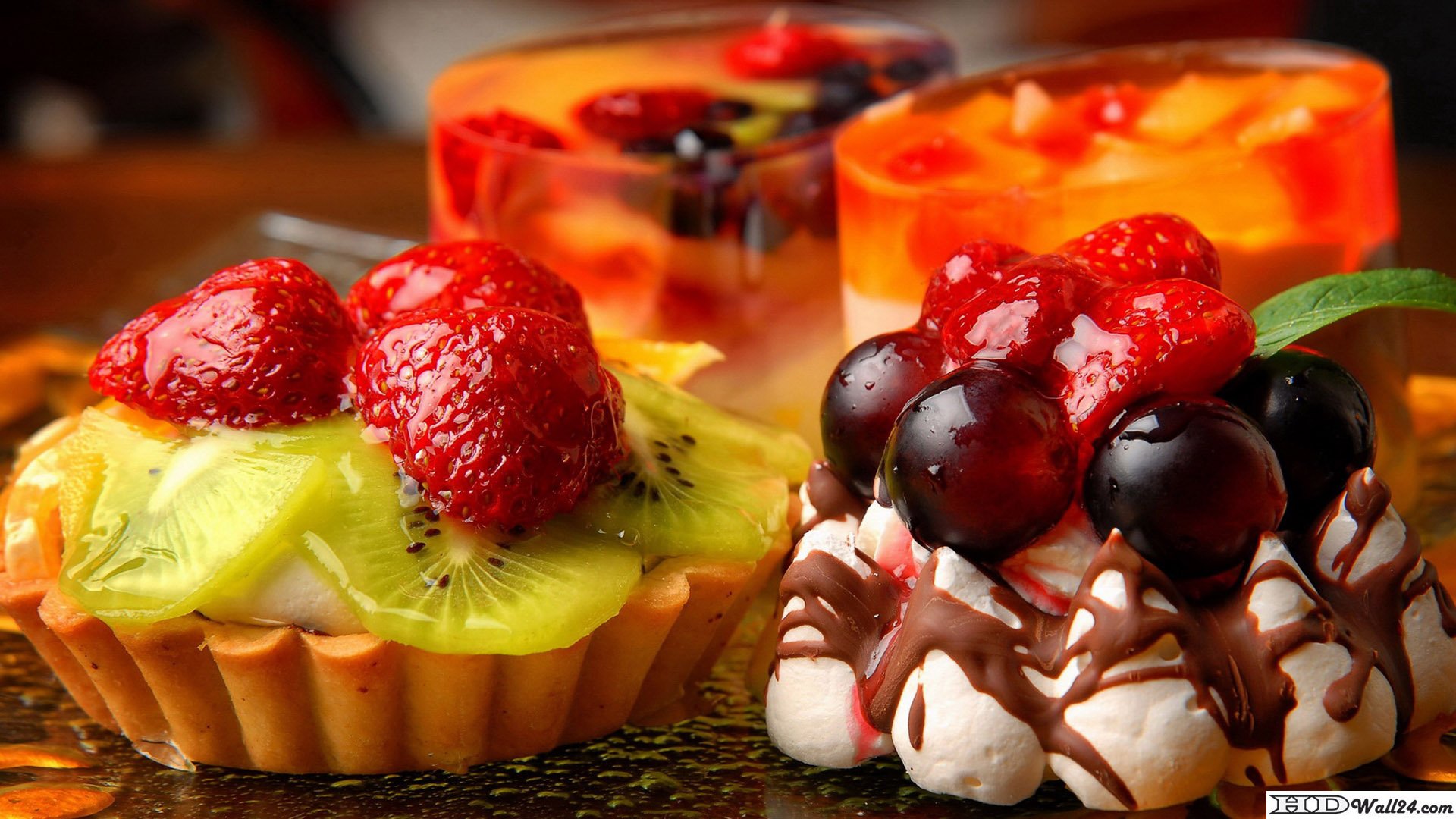HD PC desktop wallpaper of food dessert: colorful fruit tarts and chocolate-cream pastries topped with strawberries, kiwi and dark grapes, with orange jelly cups softly blurred in the background.