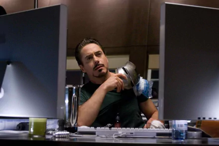 A high-definition desktop wallpaper featuring Robert Downey Jr. as Iron Man, thoughtfully engaging with technology in a modern workspace.