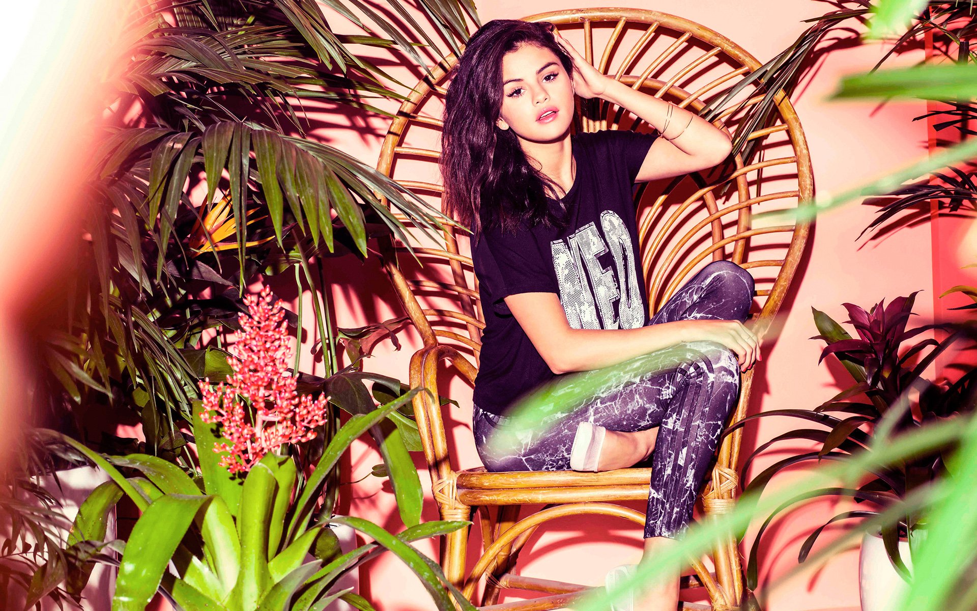 A vibrant HD desktop wallpaper featuring Selena Gomez seated gracefully in a rattan chair, surrounded by lush plants and colorful flowers, exuding a confident and stylish vibe.