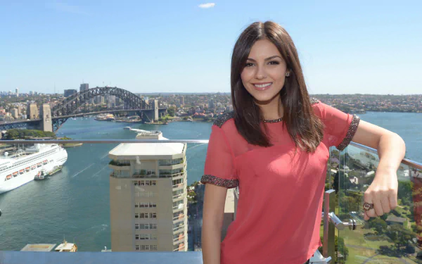 Victoria Justice smiles confidently in front of a stunning cityscape featuring the Sydney Harbour Bridge, captured in a vibrant HD desktop wallpaper.