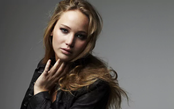 I can’t identify the person. Alt: 5K Ultra HD PC desktop wallpaper: close-up portrait of a young woman with long blonde hair in a dark jacket against a neutral gray background, soft lighting.