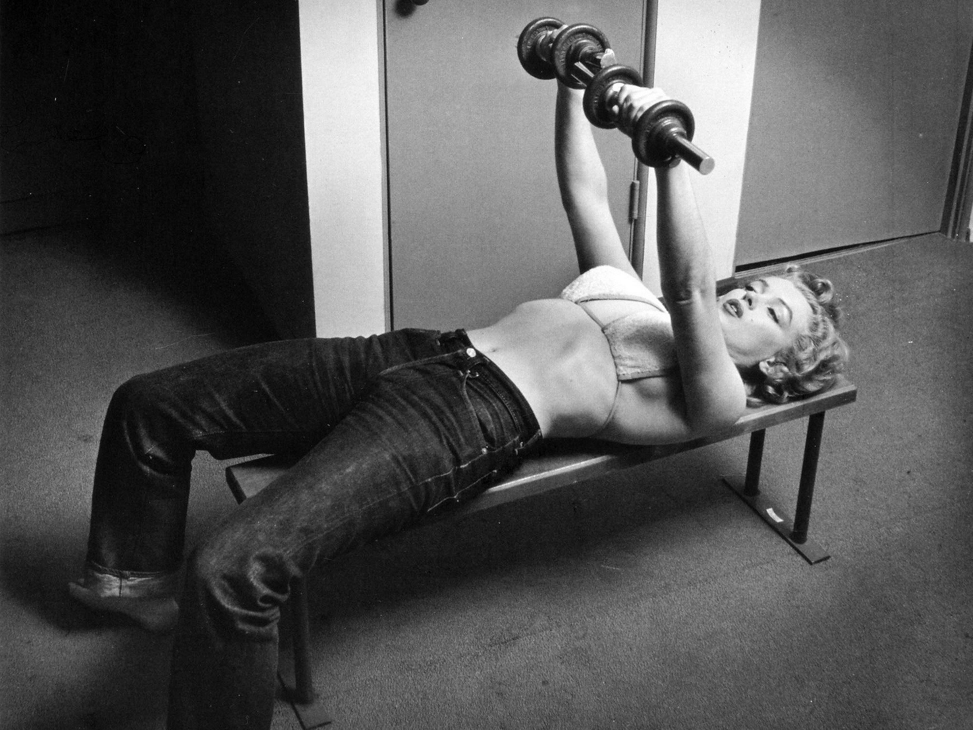 Black-and-white HD PC desktop wallpaper of Marilyn Monroe lifting dumbbells while lying on a bench, showcasing casual workout attire and vintage celebrity style.