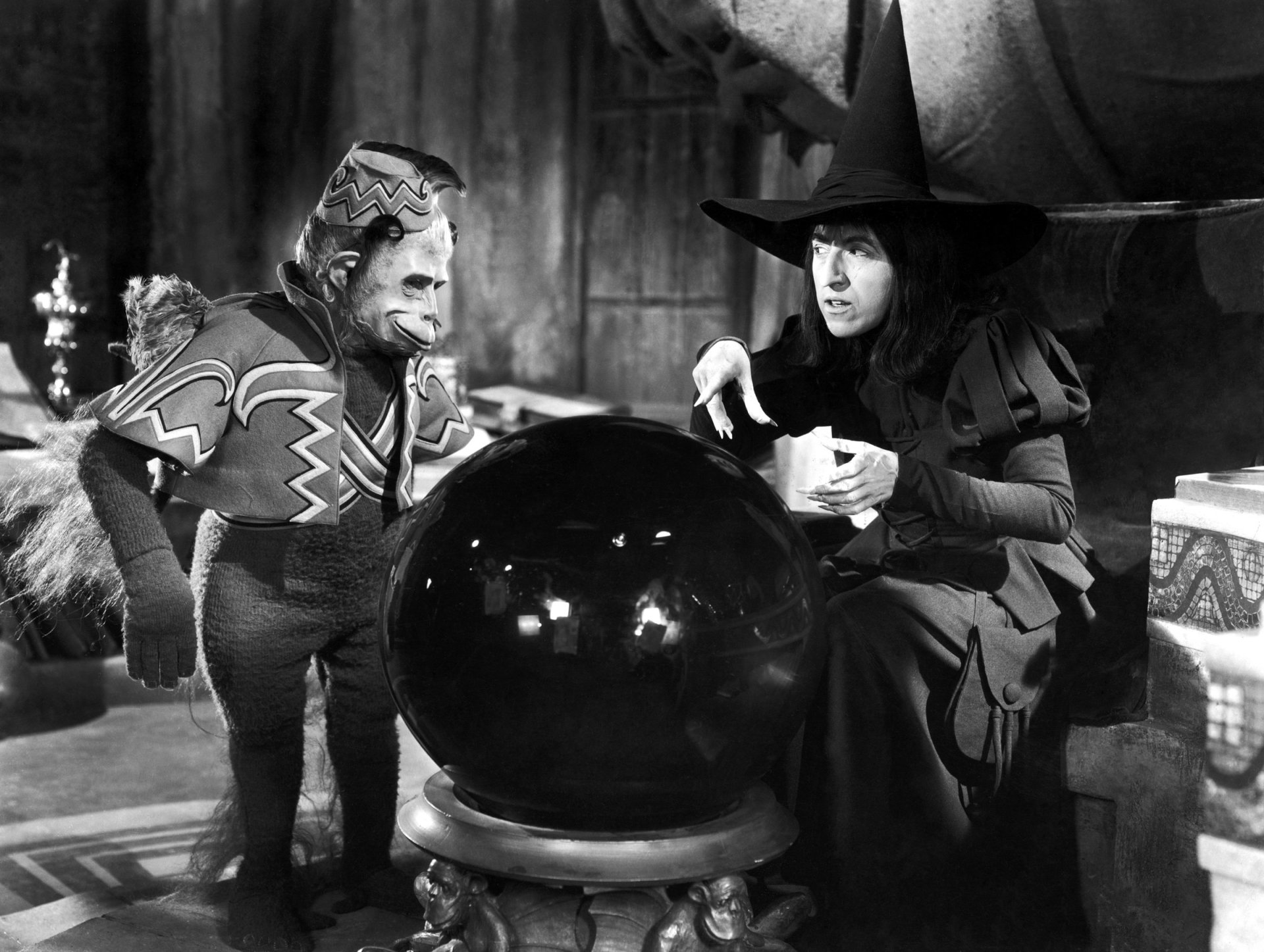 HD PC desktop wallpaper: black-and-white still from The Wizard of Oz (1939) showing the Wicked Witch of the West peering into a crystal ball while a costumed winged monkey watches.