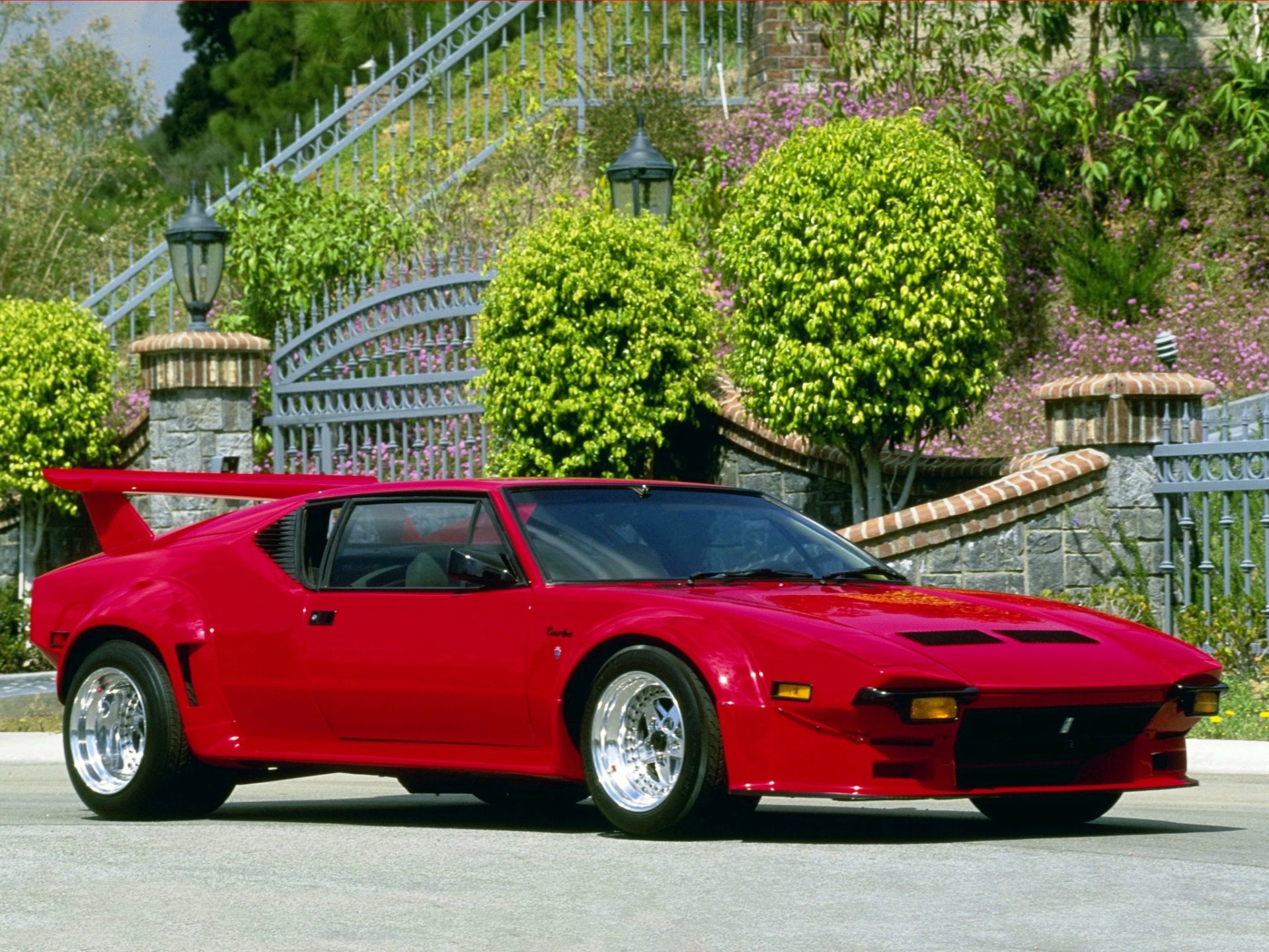 HD desktop wallpaper featuring a vibrant red De Tomaso Pantera sports car parked in front of a gated garden with trimmed bushes and stone steps.
