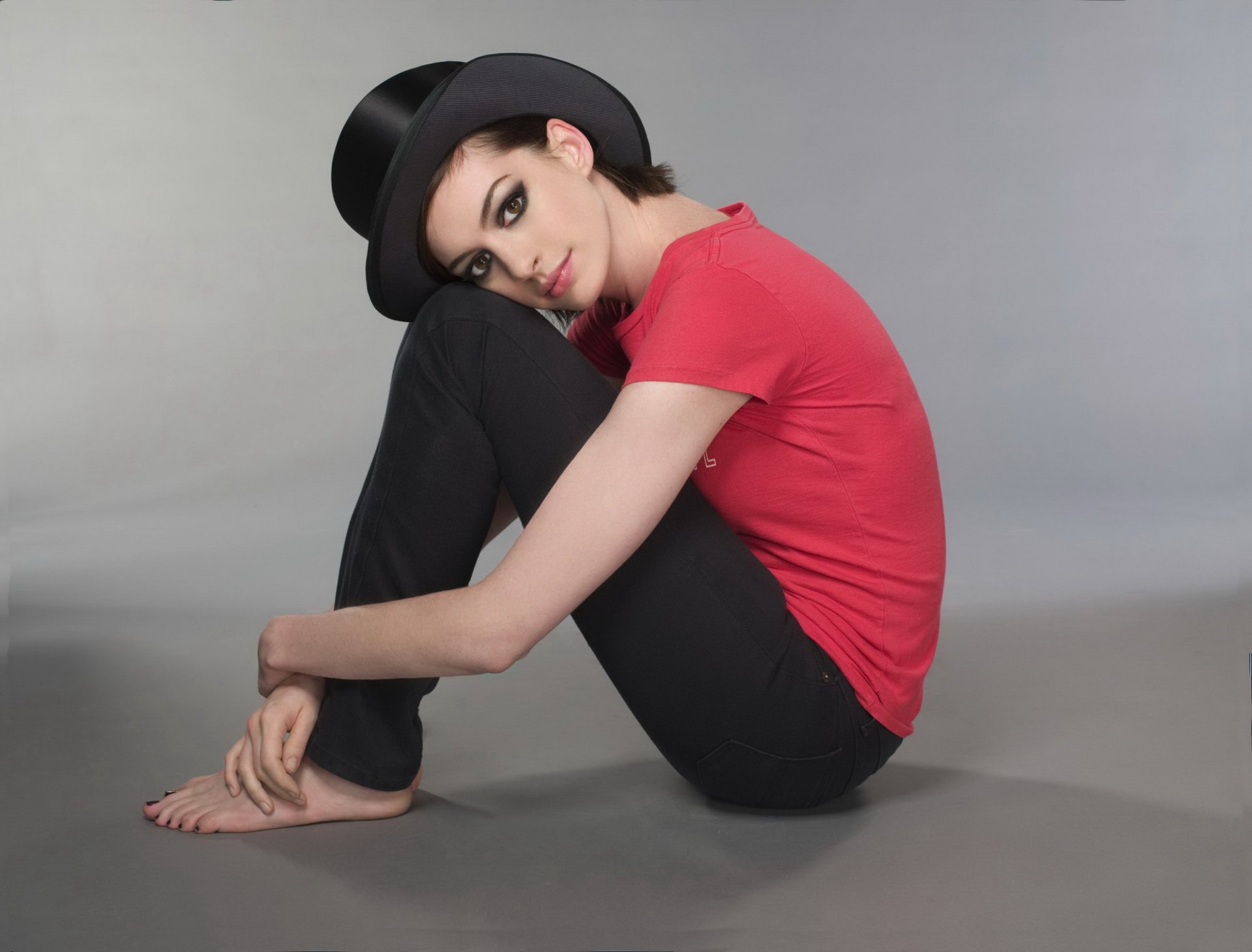 HD PC desktop wallpaper featuring celebrity Anne Hathaway sitting on the floor in a red shirt, black pants, and a black hat against a plain gray background.
