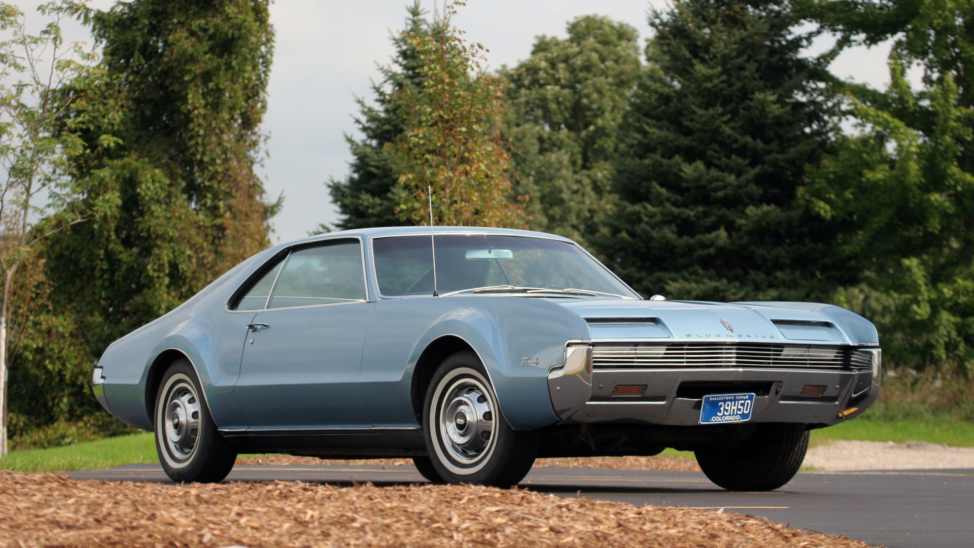 HD PC desktop wallpaper background: classic light-blue Oldsmobile Toronado vehicle parked on a tree-lined road with green foliage.