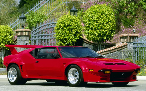 HD desktop wallpaper featuring a vibrant red De Tomaso Pantera sports car parked in front of a gated garden with trimmed bushes and stone steps.