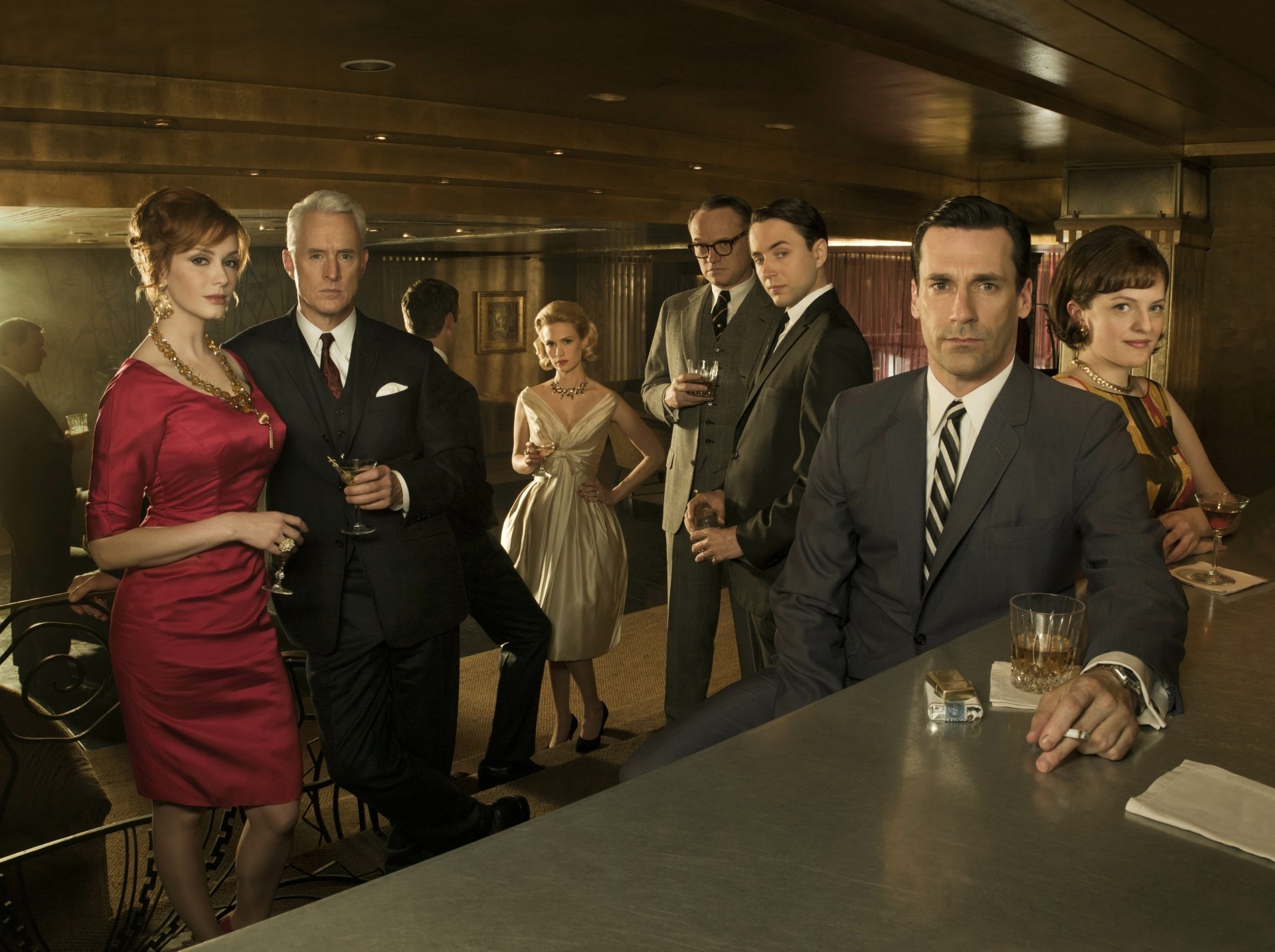 Mad Men TV show 5K Ultra HD PC desktop wallpaper/background: 1960s ad agency team in formal attire gathered at a bar, moody warm lighting and sleek mid-century office decor.
