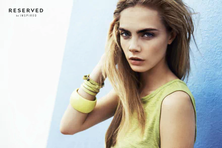 HD desktop wallpaper featuring English actress and model Cara Delevingne with blue eyes and blonde hair against a blue background, styled in a green sleeveless top.